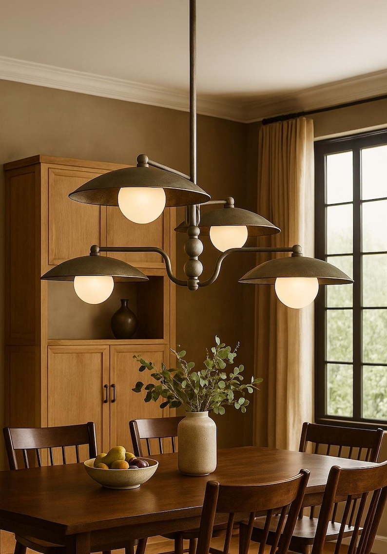 May include: A bronze-finished chandelier with four lights, each encased in a white globe, hangs above a wooden dining table. The fixture has a classic, industrial design. The dining room features a wooden cabinet, chairs, and a vase with greenery.