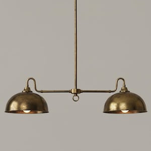 Puede incluir: Una lámpara colgante de dos luces de estilo vintage, con acabado de latón. La lámpara presenta dos pantallas en forma de cúpula, cada una con una bombilla visible. El accesorio está suspendido de una varilla central y tiene una barra horizontal que conecta las pantallas.
