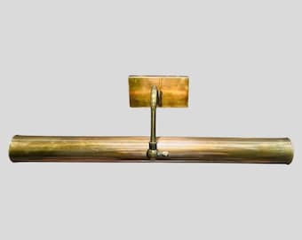 May include: A vintage brass picture light with a rectangular backplate and a long, cylindrical light fixture. The light has a curved arm connecting the backplate to the light. The brass has a warm, aged patina.