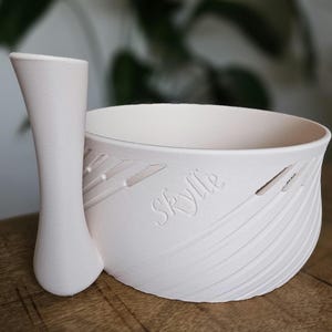 Op de afbeelding: Een witte keramische scheerkom en borstelhouder set. De kom heeft een getextureerd, gebogen ontwerp met de naam "Skyllë" op de zijkant gegraveerd. De borstelhouder is lang en slank, met een afgeronde basis.