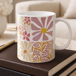 May include: White ceramic mug featuring a floral design in shades of pink, yellow, and peach. The mug sits on a stack of books, with a gold pen and glasses. A vase of flowers is in the background.