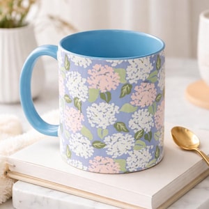 May include: A ceramic mug with a light blue handle and interior. The exterior features a floral pattern of white, pink, and green hydrangeas on a lavender background. The mug is sitting on a stack of books with a gold spoon.