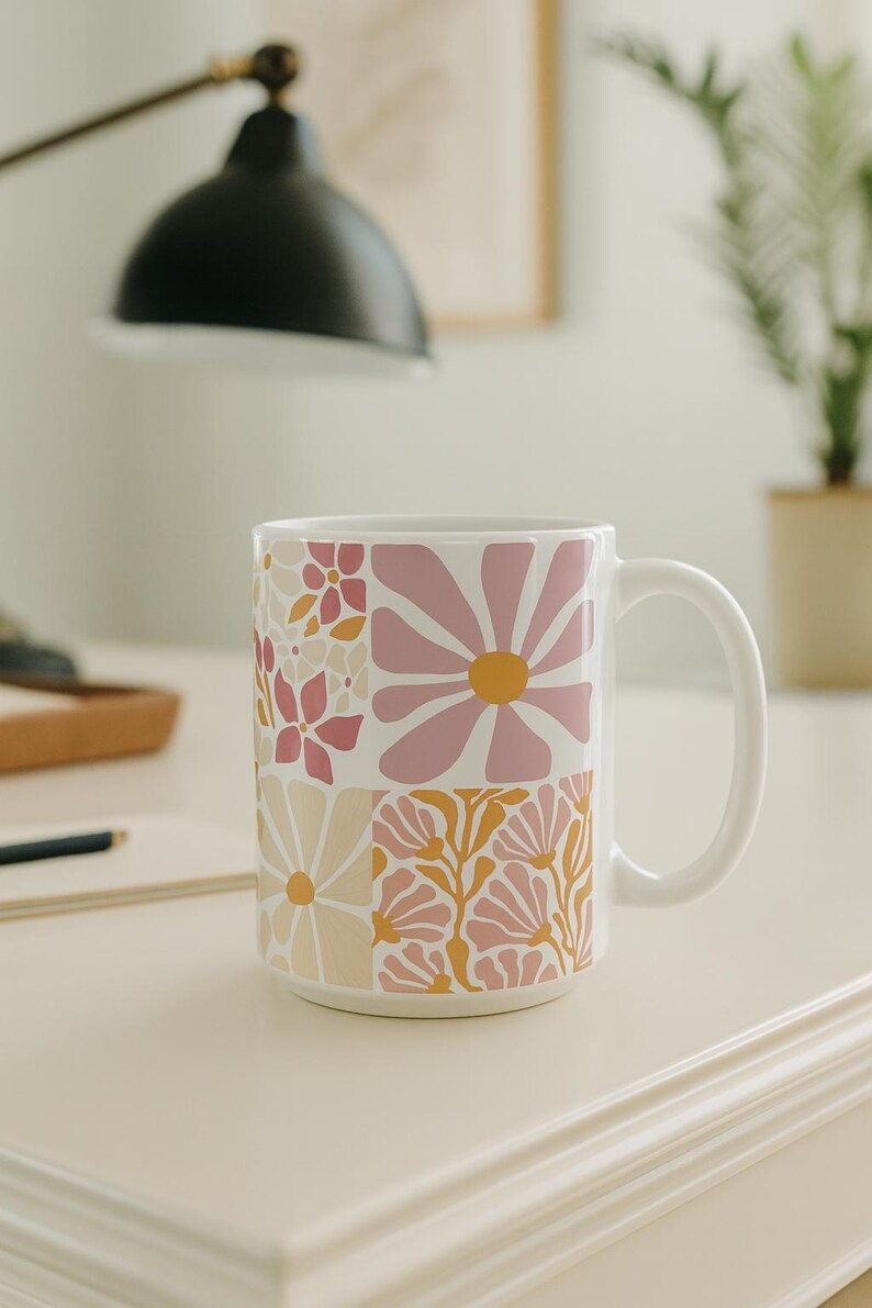 May include: White ceramic mug with a patchwork design of pink, yellow, and cream-coloured floral patterns. The mug has a large, curved handle and is set on a white surface. The background includes a desk lamp and a plant.