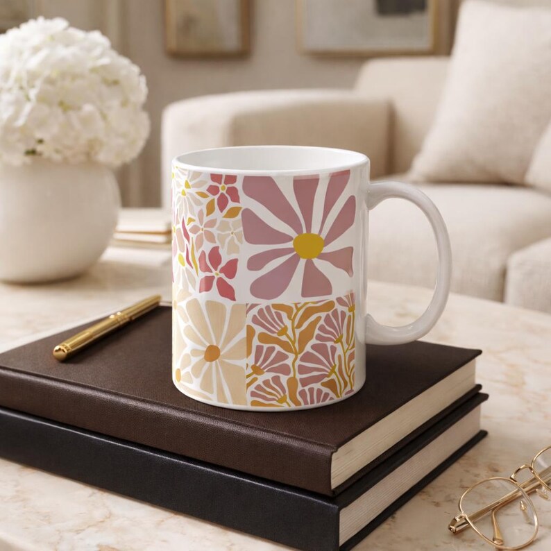 May include: White ceramic mug featuring a floral design in shades of pink, yellow, and peach. The mug sits on a stack of books, with a gold pen and glasses. A vase of flowers is in the background.