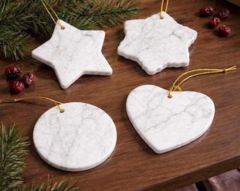 May include: Four white marble Christmas decorations in star, snowflake, circle, and heart shapes. Each has a gold string. The ornaments are on a dark wood surface with evergreen sprigs and red berries.