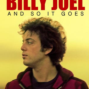 May include: Blu-ray disc cover featuring Billy Joel. The title "BILLY JOEL" is in bold red letters above the text "AND SO IT GOES". The image shows a side profile of a man with dark hair, wearing a dark shirt and a red and black plaid jacket.