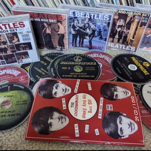 Pode incluir: Uma coleção de álbuns de música e box sets dos Beatles. A imagem mostra vários CDs e box sets com capas de álbuns com os membros da banda e os títulos dos álbuns. Os discos são pretos com texto branco, e os box sets são vermelhos e brancos.