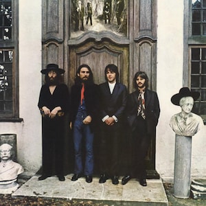 Puede incluir: Portada de álbum con cuatro hombres vestidos con trajes oscuros frente a una gran puerta de madera. Los hombres tienen el pelo largo y barba. Un busto de piedra está a la derecha. El fondo es un edificio claro.