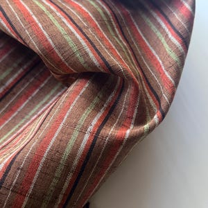 May include: A piece of fabric featuring a vertical striped pattern in shades of brown, orange, white, black, and light green. The material has a visible woven texture with a slight sheen, displayed in a folded arrangement showing the fabric's drape.