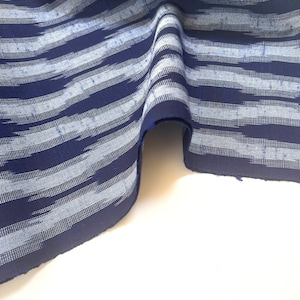 May include: Blue and white striped fabric with a subtle ikat pattern. The fabric is folded and draped on a white surface.