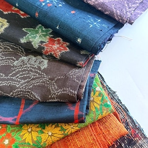 May include: A stack of colorful fabric swatches with various patterns and textures. The fabrics include floral, geometric, and abstract designs in shades of blue, orange, green, and purple. The fabrics are folded and layered, showcasing the different patterns.