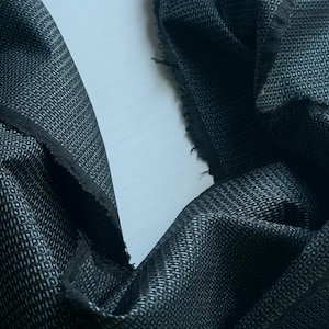 Puede incluir: Primer plano de una tela gris oscuro con un patrón sutil y texturizado. La tela está drapeada, revelando los bordes crudos y sin terminar. El tejido parece ser un pequeño diseño geométrico repetitivo, creando un aspecto sofisticado. Ideal para ropa o decoración del hogar.