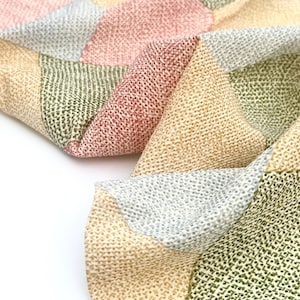 May include: Close-up of a patchwork textile with a variety of colors. The fabric features a textured pattern and is composed of geometric shapes in shades of pink, green, beige, and gray. The textile is folded, revealing its layered construction.