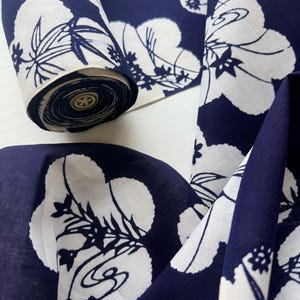 Könnte beinhalten: Marineblauer Stoff mit weißem Blumenmuster. Der Stoff ist aufgerollt, wodurch der Rand der Rolle sichtbar wird. Das Blumenmuster zeigt große, stilisierte weiße Blüten und Blätter auf einem dunkelblauen Hintergrund. Der Stoff wird wahrscheinlich für Kleidung oder Heimdekoration verwendet.