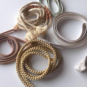 May include: A selection of decorative cords and ribbons in various colours. Includes a coiled cream cord with orange and brown accents, a gold braided cord, and ribbons in beige and green. Some cords have tassels.