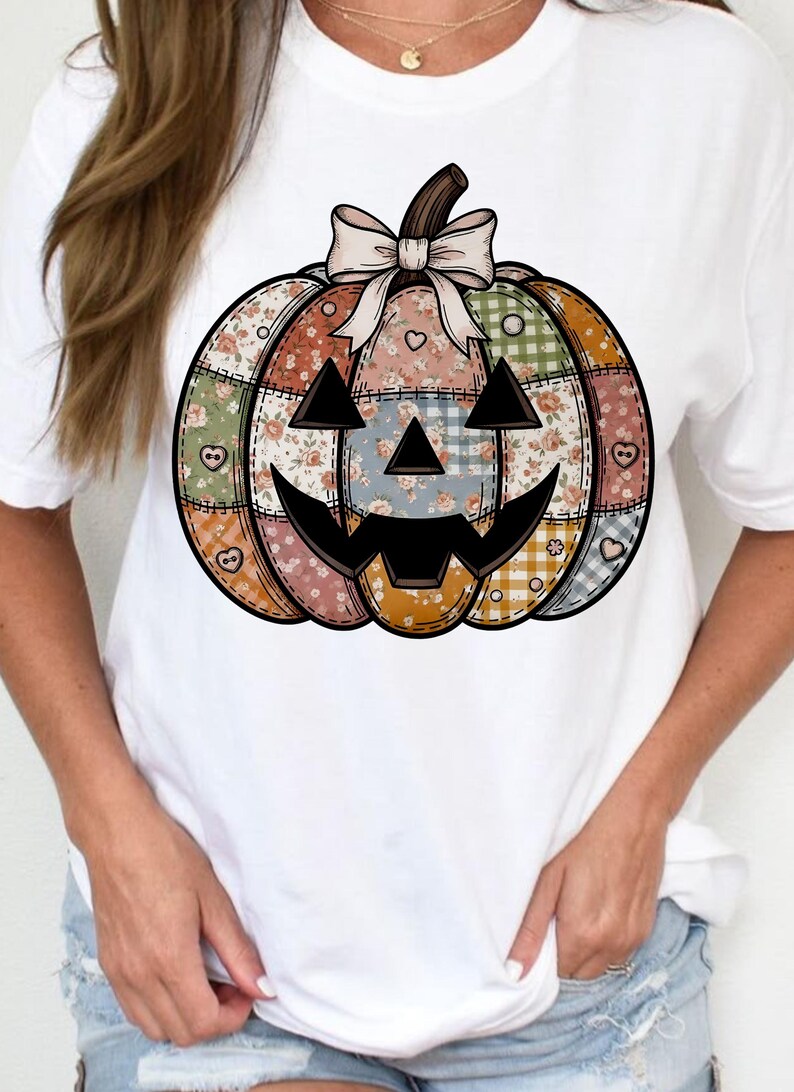 May include: White t-shirt with a patchwork pumpkin design. The pumpkin has a smiling jack-o'-lantern face and is made of various floral and patterned fabrics in orange, green, blue, and pink. A bow sits atop the pumpkin.