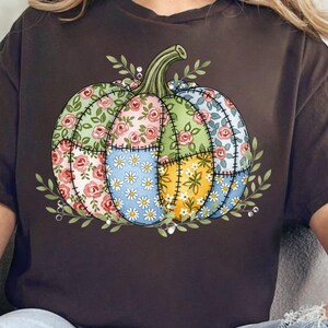 May include: A dark brown t-shirt featuring a colorful patchwork pumpkin design. The pumpkin is composed of floral patterns in pink, green, blue, and yellow, with a green stem and leaves. The design is centered on the shirt.