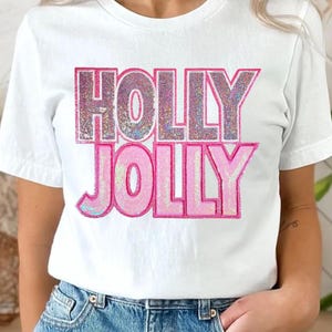 Puede incluir: Camiseta blanca con las palabras "HOLLY JOLLY" en fuente rosa con lentejuelas. La parte superior es de manga corta y la persona lleva vaqueros azules.
