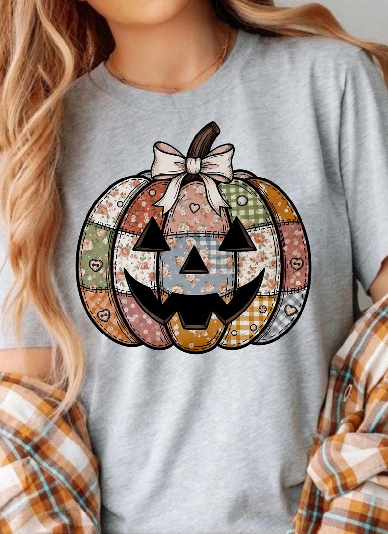 May include: A grey t-shirt with a patchwork pumpkin design. The pumpkin has a smiling jack-o'-lantern face and is decorated with floral, plaid, and heart patterns. A cream-coloured bow sits atop the pumpkin.