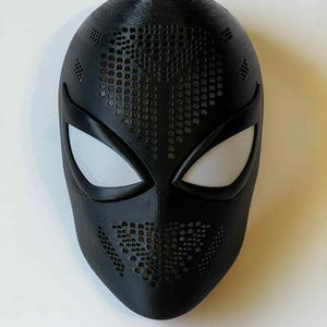 Op de afbeelding: Een zwart superheldenmasker met witte mesh oogbedekkingen. Het masker heeft een textuur en een patroon van kleine cirkelvormige perforaties over het voorhoofd en de mond. Het masker is ontworpen om op een spin te lijken.