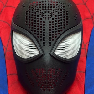 May include: A black full-face mask featuring a textured surface with numerous small circular perforations across the forehead and mouth areas. The mask includes two large, white, mesh-covered eye lenses framed in black, set against a red and blue background.