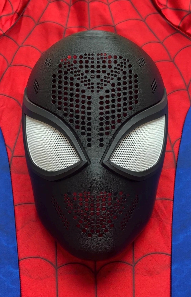 May include: A black full-face mask featuring a textured surface with numerous small circular perforations across the forehead and mouth areas. The mask includes two large, white, mesh-covered eye lenses framed in black, set against a red and blue background.