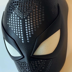 May include: A black superhero mask with white mesh eye coverings and a textured surface. The mask features a pattern of small, evenly spaced holes across the forehead and cheeks, and is likely a costume accessory.