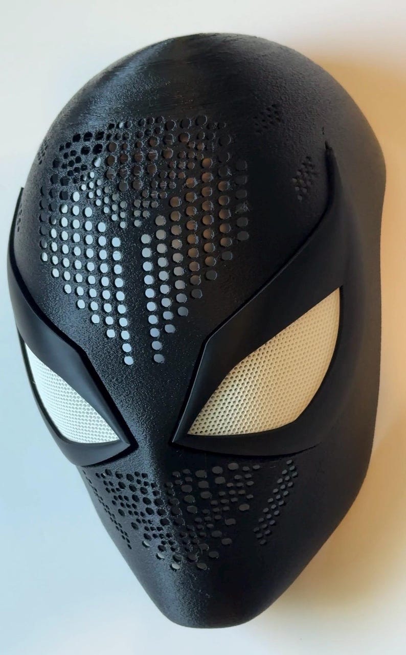 May include: A black superhero mask with white mesh eye coverings and a textured surface. The mask features a pattern of small, evenly spaced holes across the forehead and cheeks, and is likely a costume accessory.