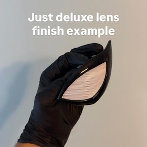 Op de afbeelding: Zwarte Spiderman-stijl ooglens met een wit mesh-patroon. De tekst "Just deluxe lens finish example" wordt in het wit weergegeven. De lens wordt vastgehouden door een gehandschoende hand.