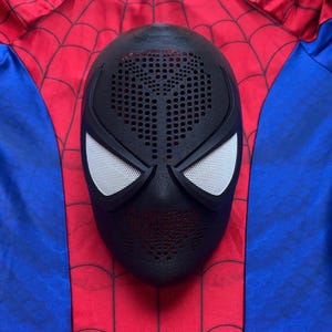 May include: A black full-face mask featuring a textured surface with numerous small circular perforations across the forehead and chin areas, paired with two large, white, mesh-covered eye lenses, resting on a red and blue fabric background.
