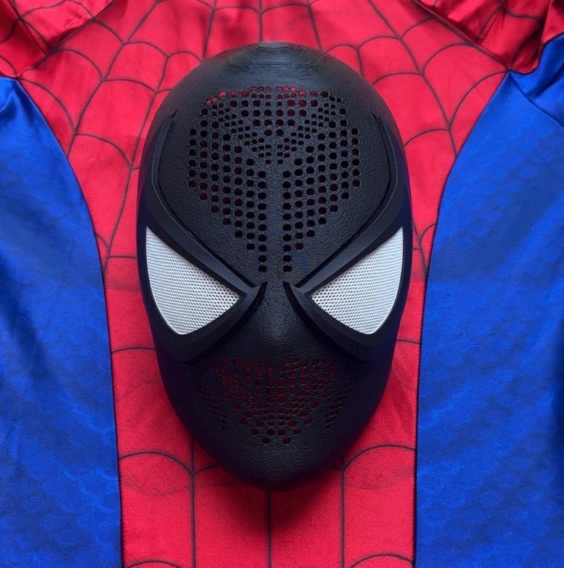 May include: A black full-face mask featuring a textured surface with numerous small circular perforations across the forehead and chin areas, paired with two large, white, mesh-covered eye lenses, resting on a red and blue fabric background.