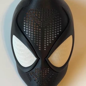 Op de afbeelding: Een zwart superheldenmasker met witte mesh oogbedekkingen. Het masker heeft een getextureerd oppervlak en een patroon van kleine gaatjes rond de mond en het voorhoofd. Het masker is waarschijnlijk voor cosplay of kostuumgebruik.