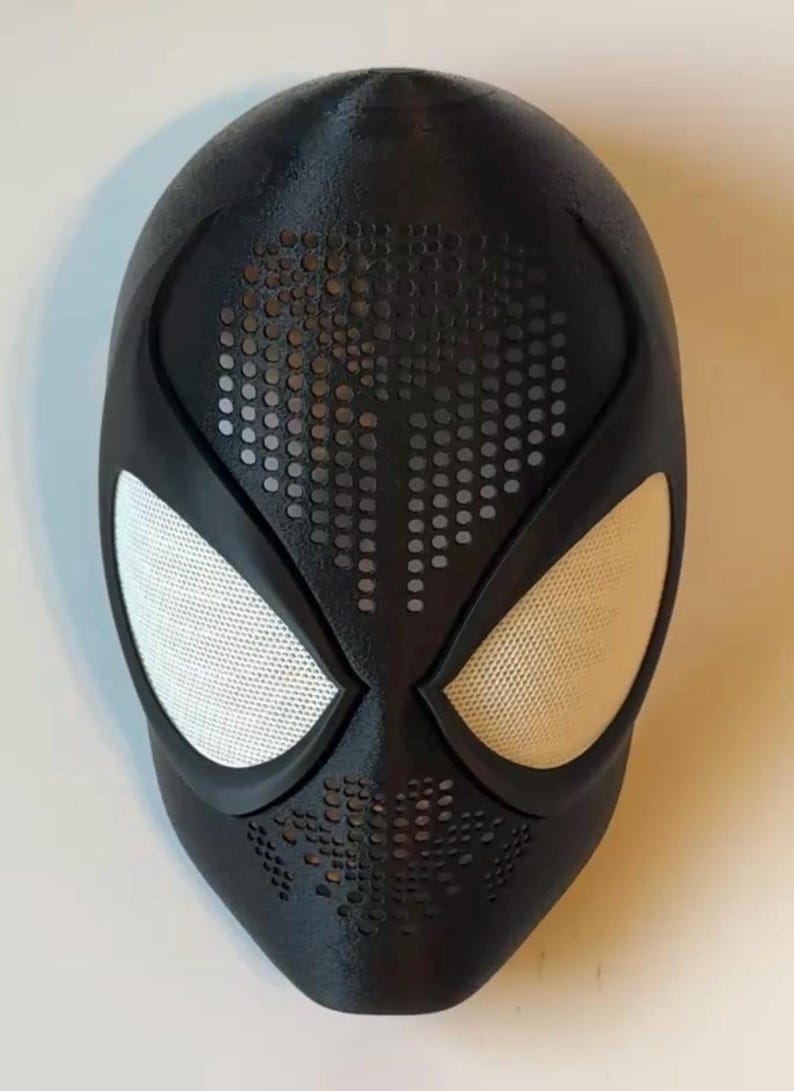 Op de afbeelding: Een zwart superheldenmasker met witte mesh oogbedekkingen. Het masker heeft een getextureerd oppervlak en een patroon van kleine gaatjes rond de mond en het voorhoofd. Het masker is waarschijnlijk voor cosplay of kostuumgebruik.