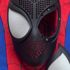 May include: A black mask with a textured, perforated surface and a large eye opening, featuring a removable white mesh eye lens. The mask is positioned over a red and blue fabric background with a black spiderweb pattern printed on the red sections.