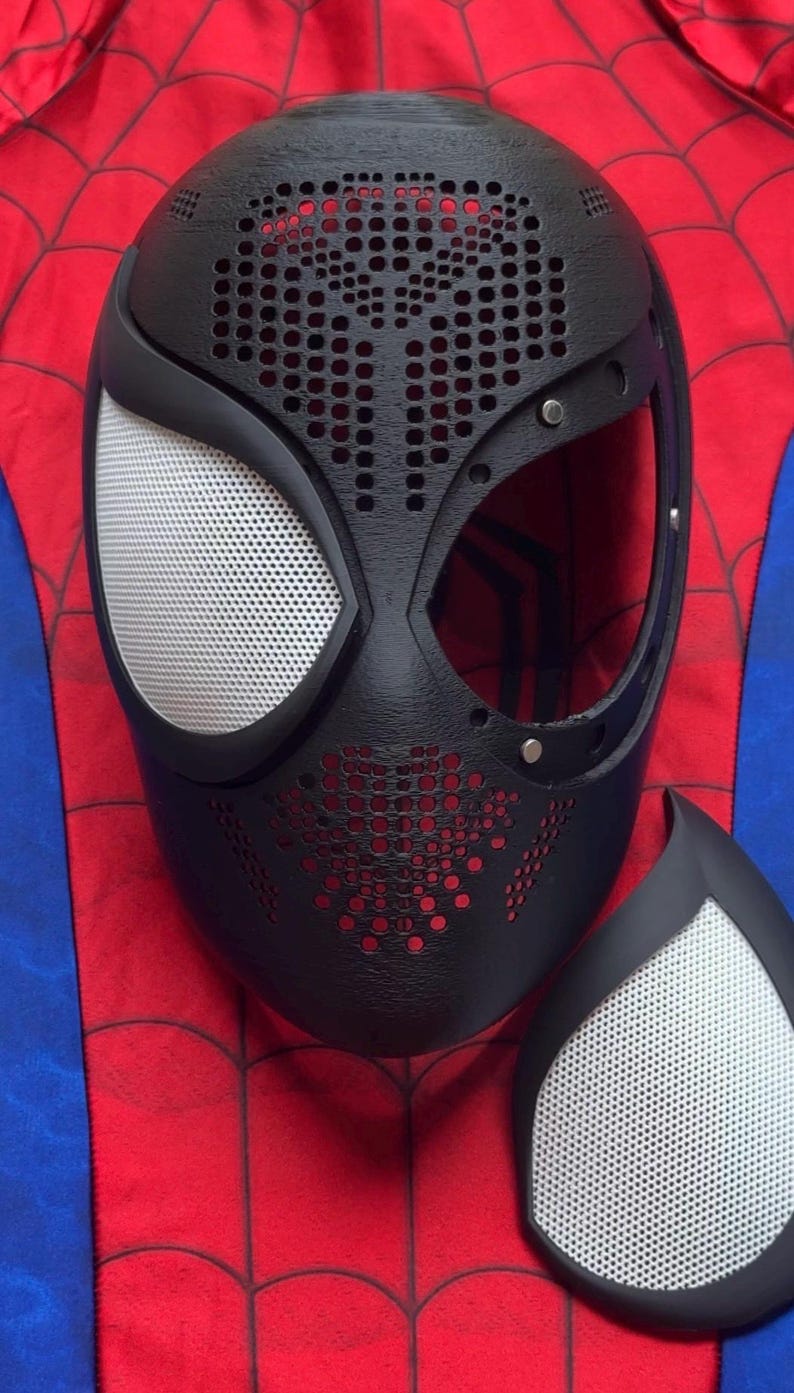 May include: A black mask with a textured, perforated surface and a large eye opening, featuring a removable white mesh eye lens. The mask is positioned over a red and blue fabric background with a black spiderweb pattern printed on the red sections.