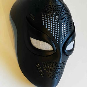 Puede incluir: Una m&aacute;scara negra con una superficie texturizada y aberturas para los ojos cubiertas con malla blanca. La m&aacute;scara tiene una serie de peque&ntilde;os agujeros en la parte superior y los lados. La m&aacute;scara es probablemente para cosplay o disfraces.