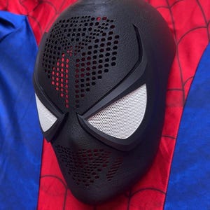 May include: A black full-face mask with a textured surface, featuring a perforated pattern across the forehead and mouth area. The mask includes two large, white, mesh-covered eye lenses framed by thick black borders, designed for a costume.