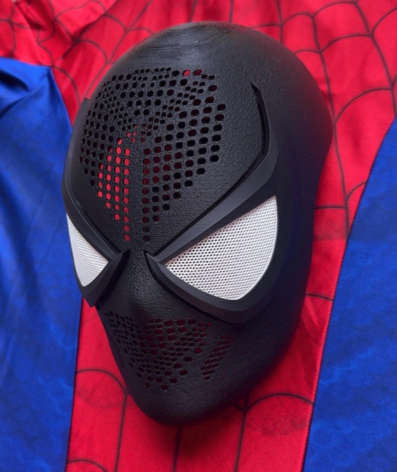 May include: A black full-face mask with a textured surface, featuring a perforated pattern across the forehead and mouth area. The mask includes two large, white, mesh-covered eye lenses framed by thick black borders, designed for a costume.