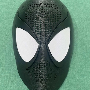 Op de afbeelding: Een zwart masker met witte oogbedekkingen, dat lijkt op een superheldenmasker. Het masker heeft een textuuroppervlak met kleine perforaties aan de bovenkant. De oogbedekkingen zijn wit en passen bij het ontwerp van het masker.