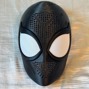Op de afbeelding: Een zwart masker met witte mesh oogbedekkingen. Het masker heeft een gestructureerd oppervlak met kleine cirkelvormige perforaties. De oogopeningen zijn zwart omlijnd. Accessoire voor een superhelden kostuum.