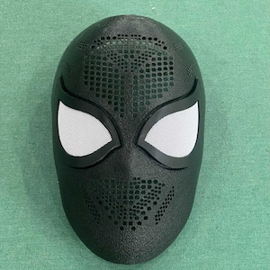 May include: Black superhero mask with white mesh eye coverings and a perforated design. The mask is a full face covering, likely for cosplay or costume use. The mask has a textured surface.