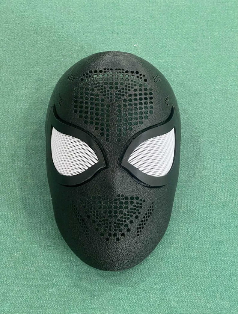 May include: Black superhero mask with white mesh eye coverings and a perforated design. The mask is a full face covering, likely for cosplay or costume use. The mask has a textured surface.
