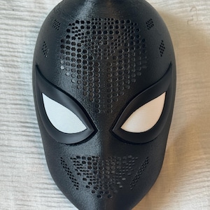 以下が含まれることがあります： 小さな穴のデザインとテクスチャ加工された表面を持つ黒いマスク。マスクには白いメッシュの目の覆いと黒い目の周囲があります。マスクはおそらくコスプレまたはコスチューム用です。