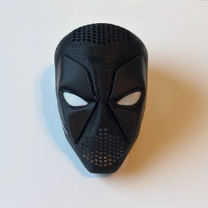 May include: A black mask with a textured surface, featuring a design with white mesh eye openings and a perforated mouth area. The mask has a distinctive shape with angular features and is likely for cosplay or costume use.