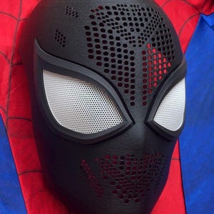 May include: A black full-face mask featuring a textured surface with a perforated pattern across the forehead and chin areas. The mask includes two large, almond-shaped eye openings covered with a white mesh material and framed in black.