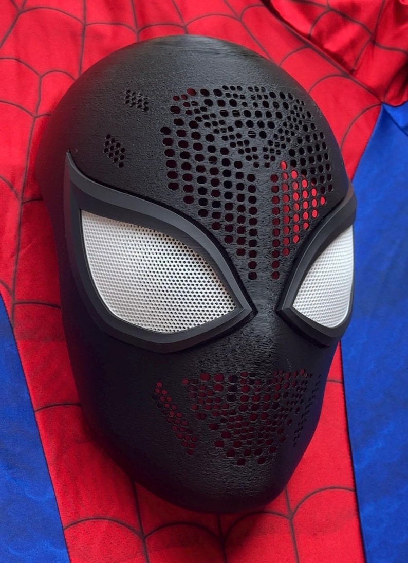 May include: A black full-face mask featuring a textured surface with a perforated pattern across the forehead and chin areas. The mask includes two large, almond-shaped eye openings covered with a white mesh material and framed in black.