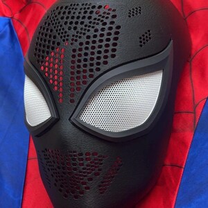 May include: A black full-face mask featuring a textured surface with numerous small circular perforations across the forehead and chin areas. The mask includes two large, almond-shaped eye openings covered with a fine white mesh material.