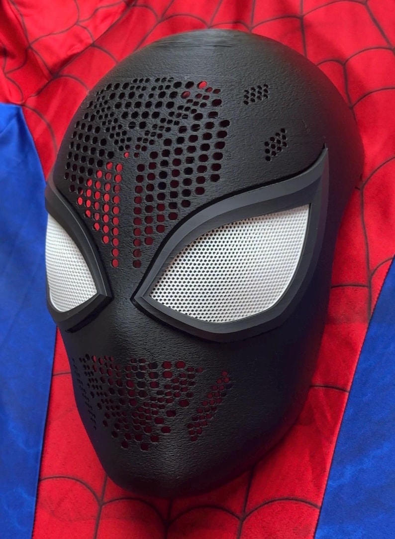 May include: A black full-face mask featuring a textured surface with numerous small circular perforations across the forehead and chin areas. The mask includes two large, almond-shaped eye openings covered with a fine white mesh material.
