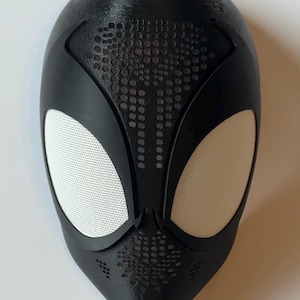 May include: Black superhero mask with white mesh eye coverings. The mask has a textured surface and a series of small, evenly spaced holes on the forehead and around the mouth area. The mask is designed to cover the entire face.