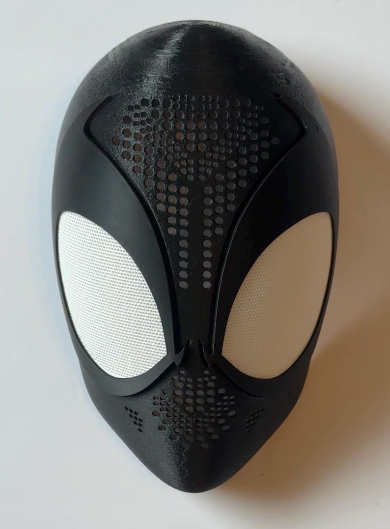 May include: Black superhero mask with white mesh eye coverings. The mask has a textured surface and a series of small, evenly spaced holes on the forehead and around the mouth area. The mask is designed to cover the entire face.